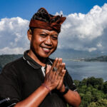 A man wearing traditional Balinese headwear presses his palms together in greeting, with a lake and hills behind him