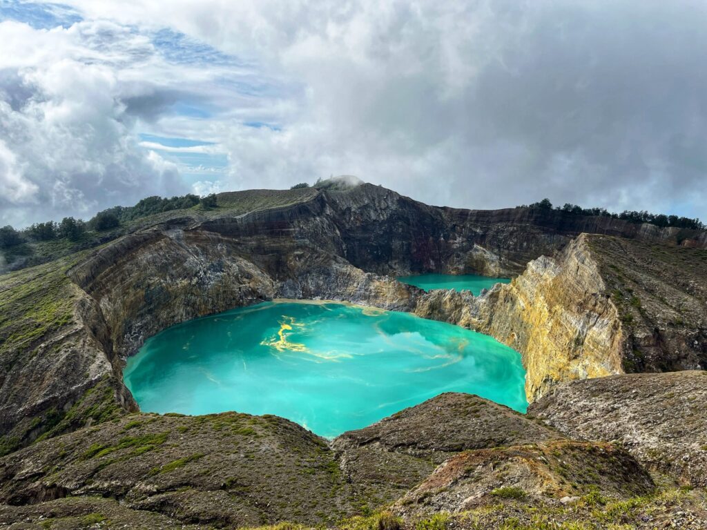 Turquoise volcanic crater lake surrounded by rocky ridges, with a second lake visible beyond under a cloudy sky