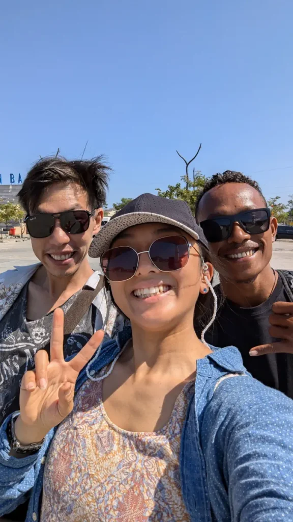 Three smiling people take a selfie outdoors in sunglasses and a cap, one flashing a peace sign, under a clear blue sky