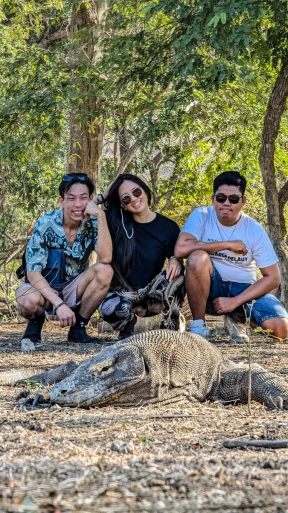 Three people crouch smiling behind a large Komodo dragon resting on the ground in a wooded area