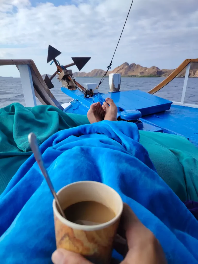 POV shot of bare feet resting on a boat's blue bow deck, holding a cup of coffee, with hazy mountains across the water