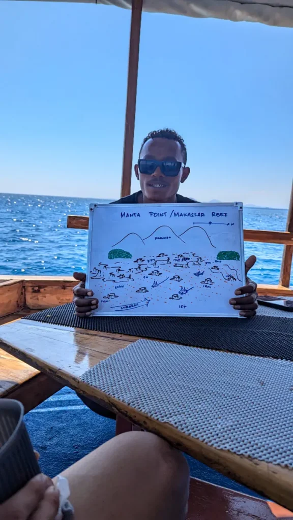 Man in sunglasses aboard a boat holds up a hand-drawn whiteboard dive site map, ocean visible behind him
