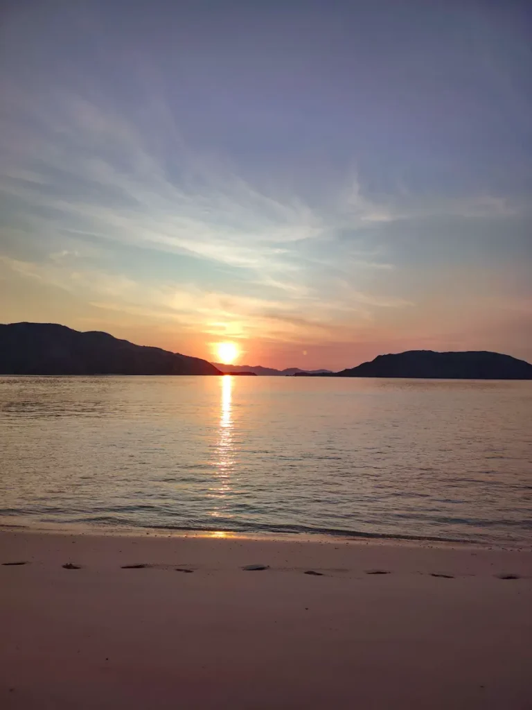 Sun setting over calm water between two dark hillsides, with footprints in the sand along the shoreline in the foreground
