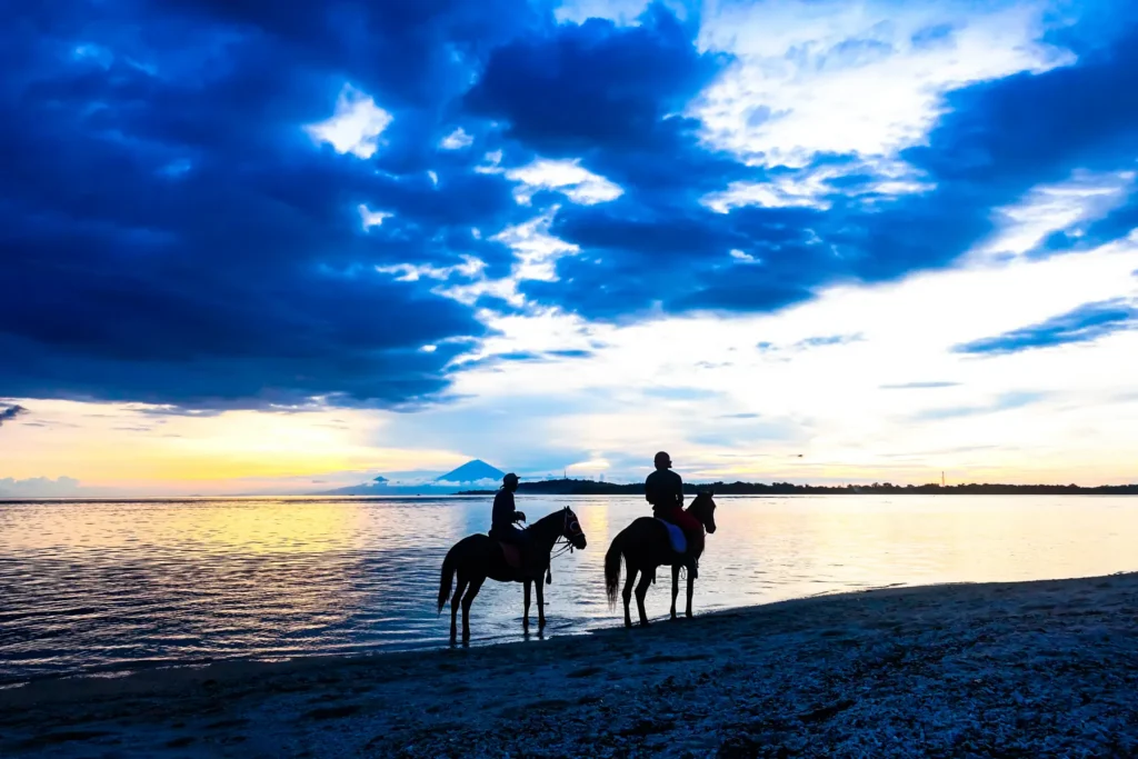 Deux cavaliers silhouettés sur une plage au bord de l'eau, sous un ciel nuageux au coucher du soleil avec un volcan au loin
