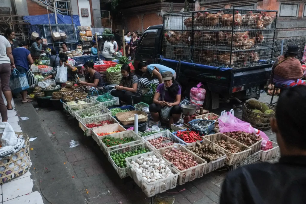 A busy street market with vendors selling produce from crates on the ground beside a truck carrying caged chickens