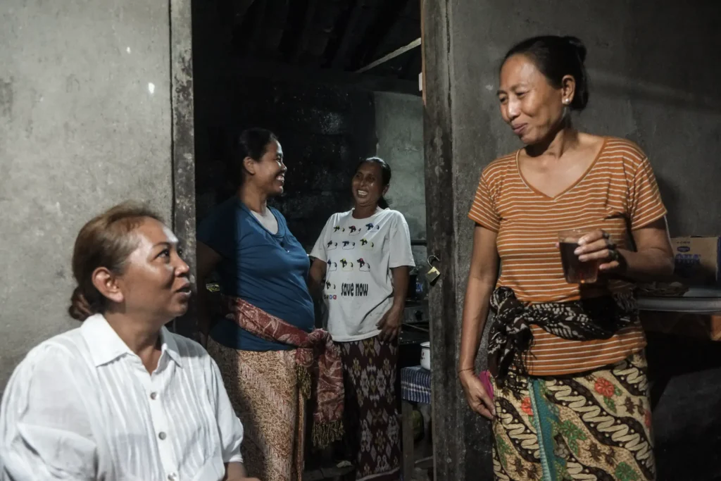 Four women gather and smile inside a modest home, one holding a glass, wearing patterned sarongs and casual tops