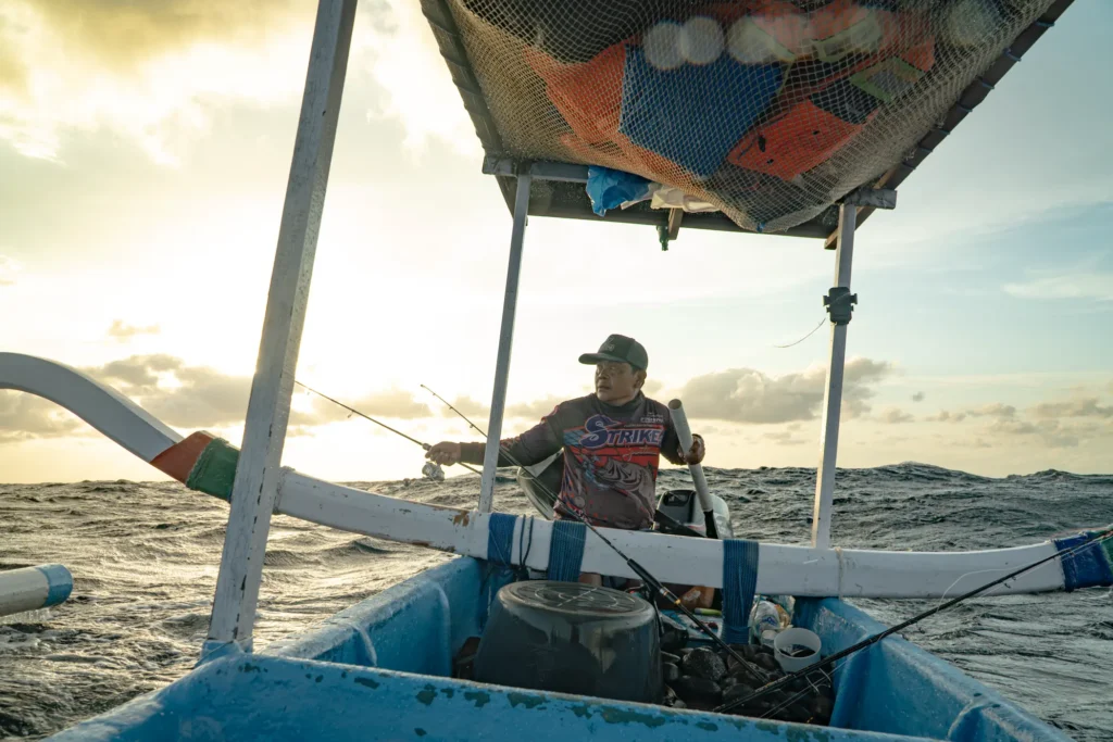 A fisherman in a cap holds a rod aboard an outrigger boat with a canopy, ocean and sunrise sky behind him