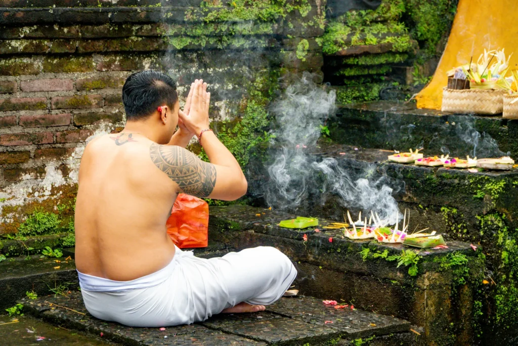 A shirtless man with a tattooed arm sits cross-legged in prayer position before smoking incense and offerings placed on stone steps