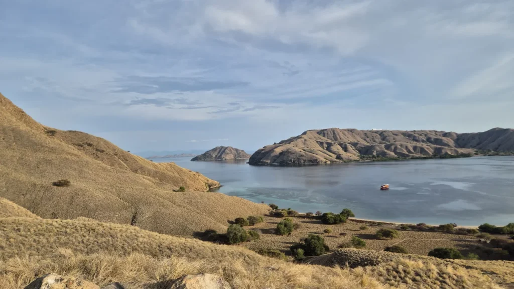 Dry grassy hills overlook a calm bay with a small boat anchored near distant coastal cliffs under a hazy sky