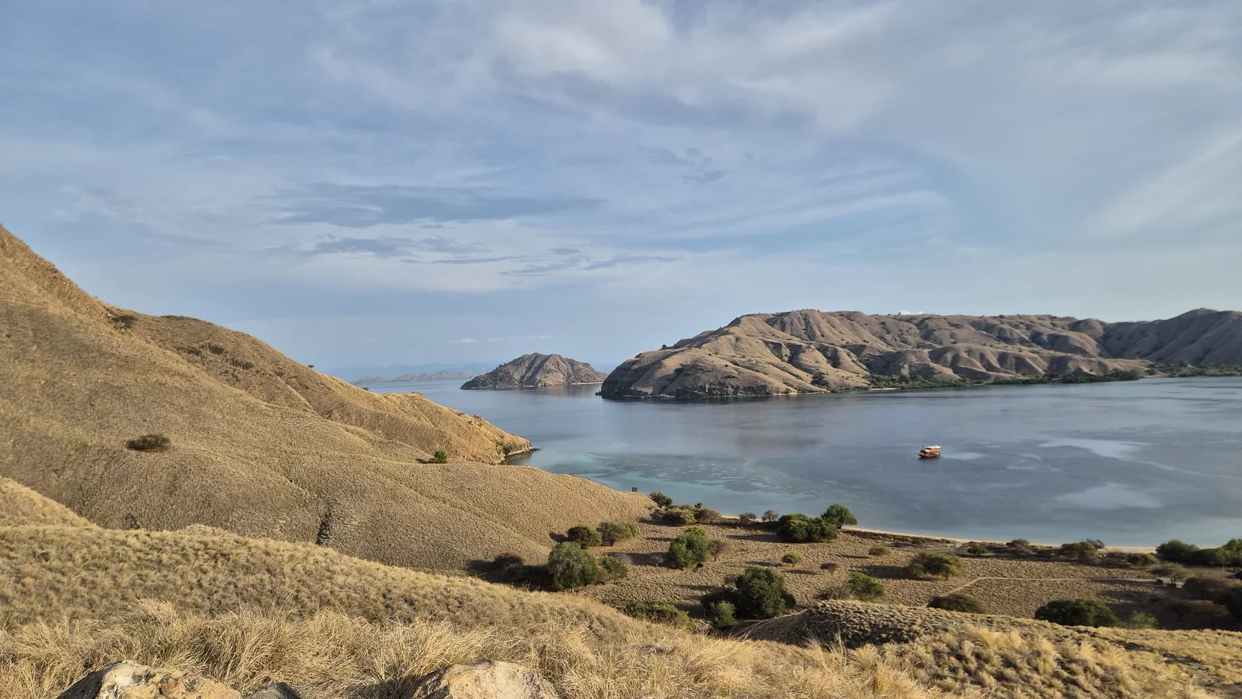 Collines arides surplombant une baie calme où mouille un petit bateau, entre plage claire et végétation clairsemée