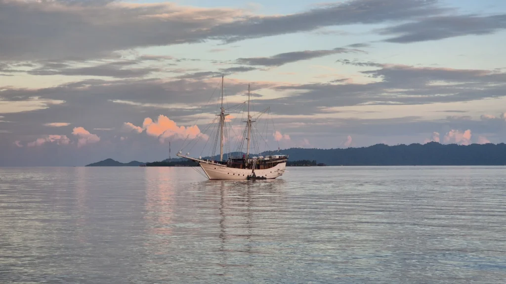A twin-masted wooden sailing boat anchored on calm water at dusk, with distant hills silhouetted under a pastel cloudy sky