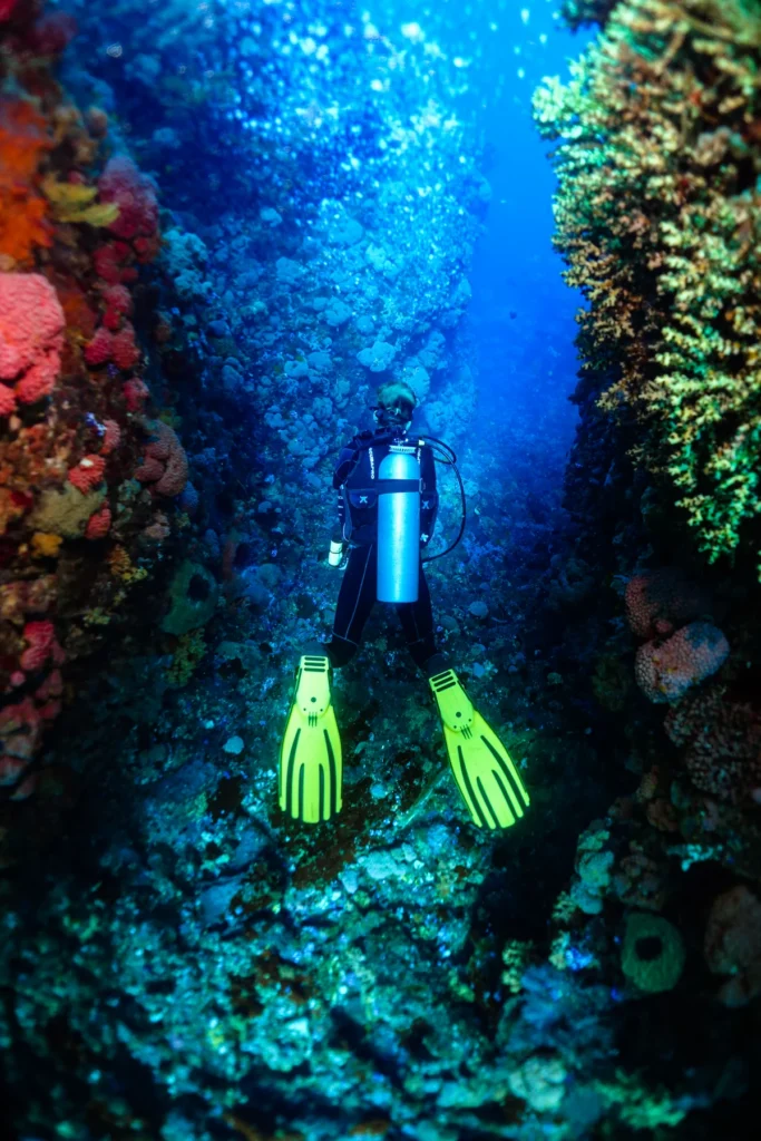 A scuba diver with bright green fins swims through a narrow crevice between two coral-covered rock walls