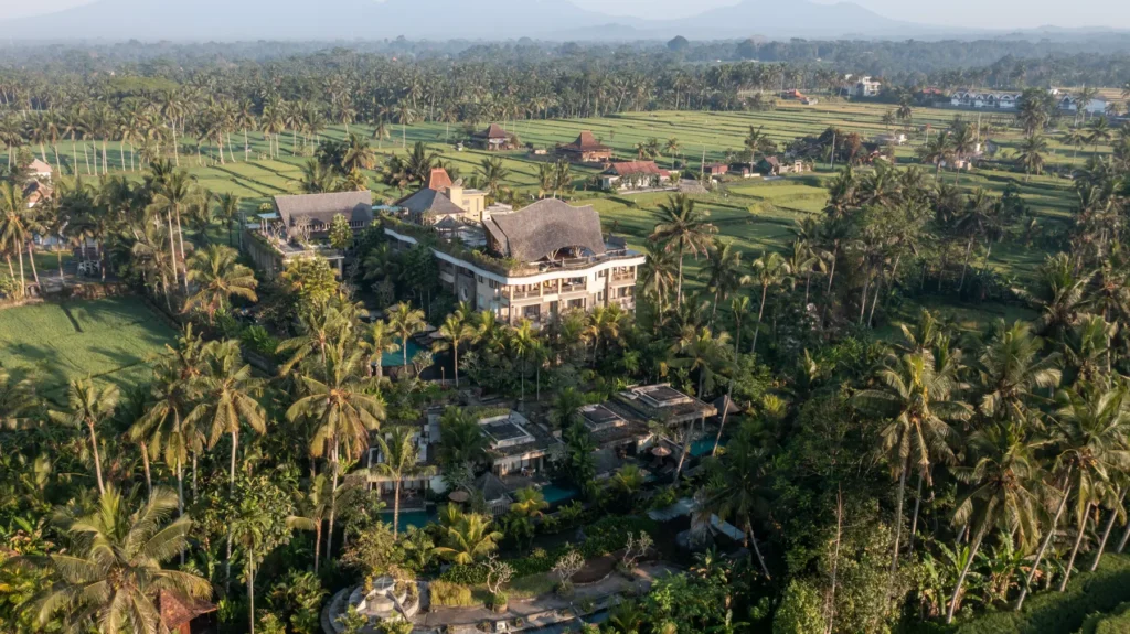 Aerial view of Kuwarasan Ubud's main building with thatched roof, surrounded by palm trees, gardens and rice fields extending into the distance
