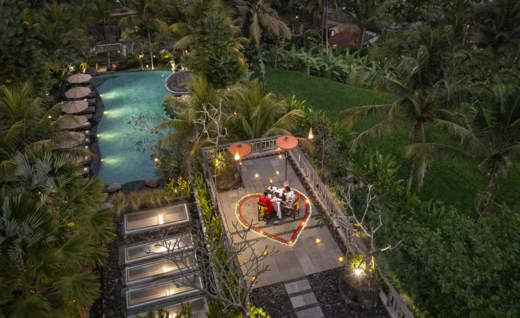 Aerial view of Kuwarasan Ubud's grounds at night showing an illuminated pool, a terrace with gathered guests, tropical vegetation, and pathway lighting