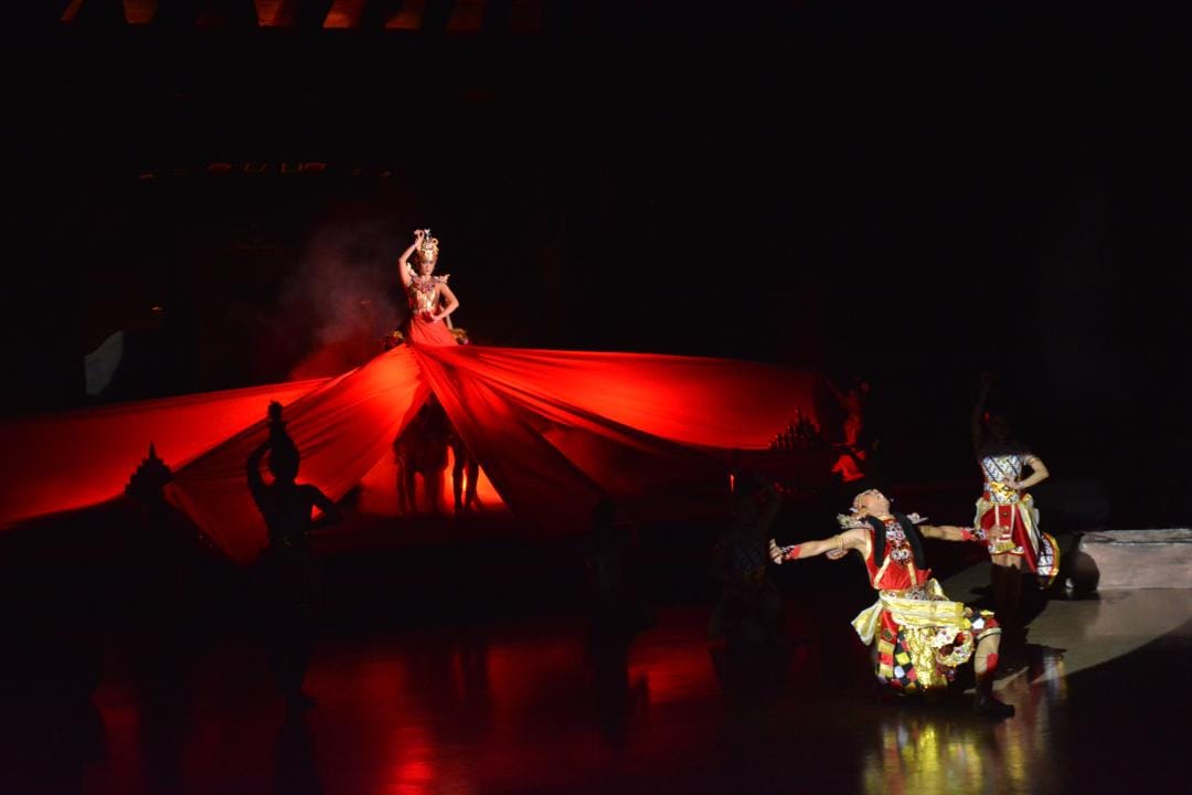 A stage performer in an ornate red gown atop a dramatic flowing red fabric backdrop, with dancers in patterned costumes in the foreground