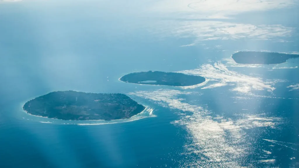 Aerial view of three green islands scattered across a blue sea with sunlight reflecting off scattered clouds and water