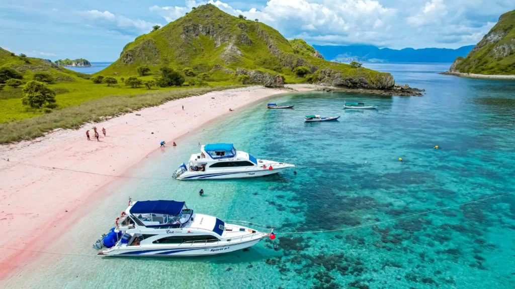 Aerial view of two boats anchored in turquoise water beside a pink-sand beach with a green hill and small islands in the distance