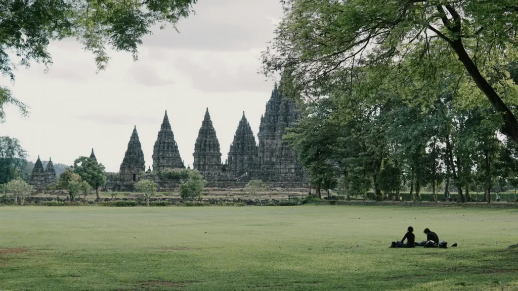 Distant stone temple spires rise beyond a wide grassy field where two people sit in the shade of trees