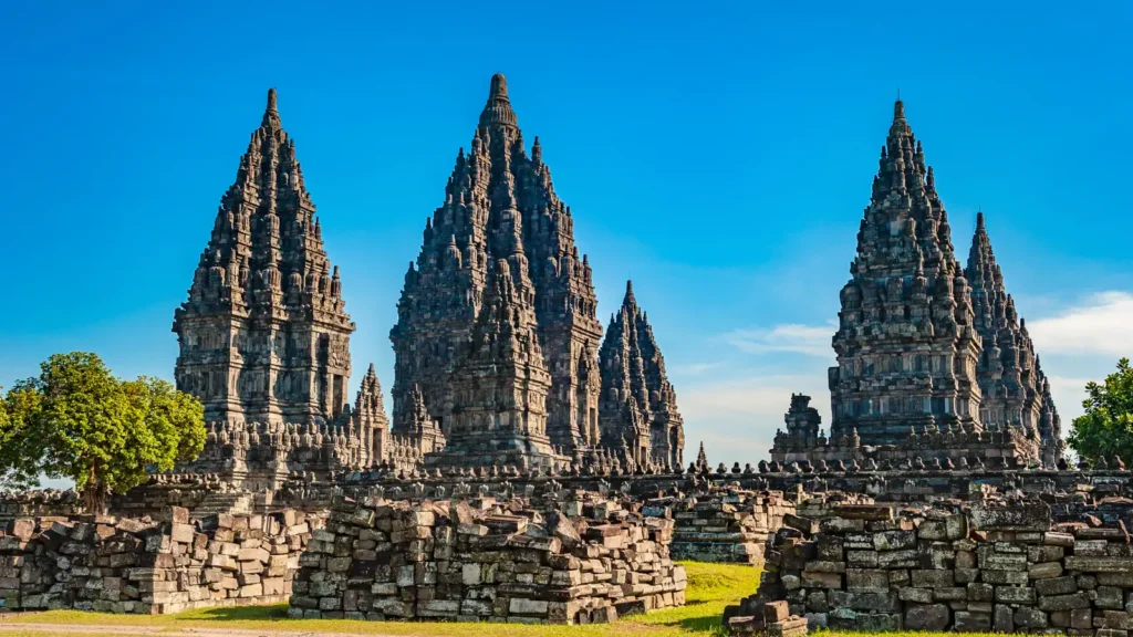 Tall stone temple spires with intricately carved tiers rise against a blue sky, with scattered ruins and a tree in the foreground