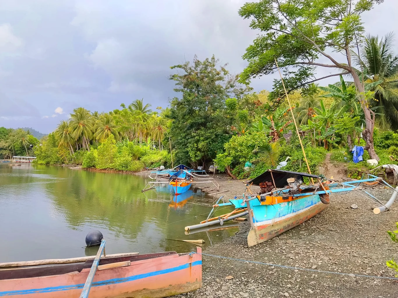 Traditional outrigger boats rest on a gravel riverbank lined with palm trees and dense green vegetation under a cloudy sky