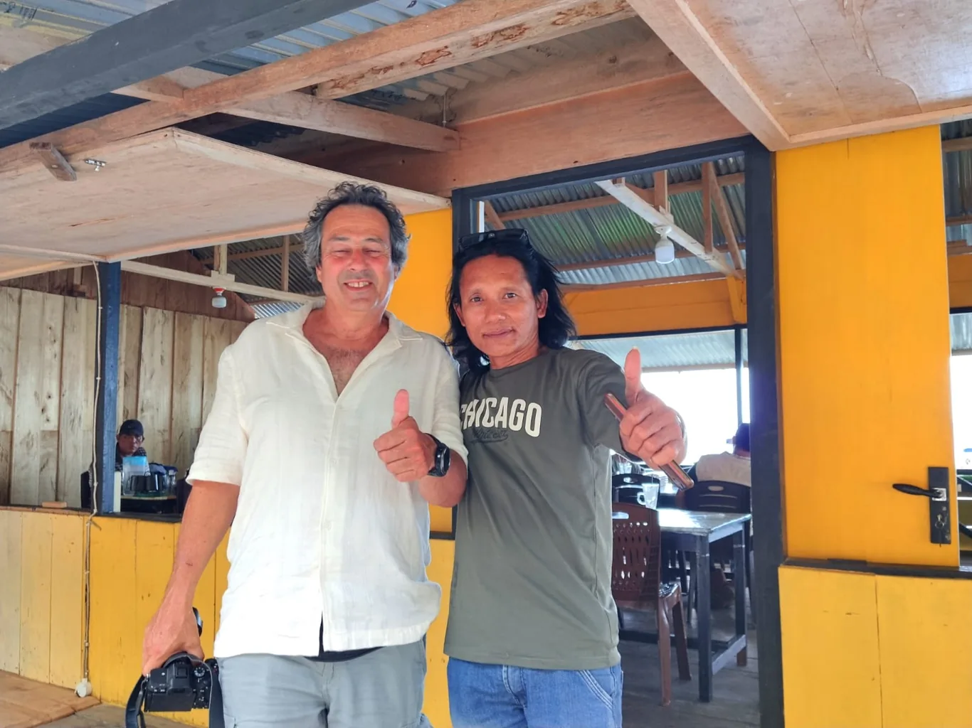 Two men give thumbs up together under a covered wooden shelter with yellow support beams and corrugated metal roofing