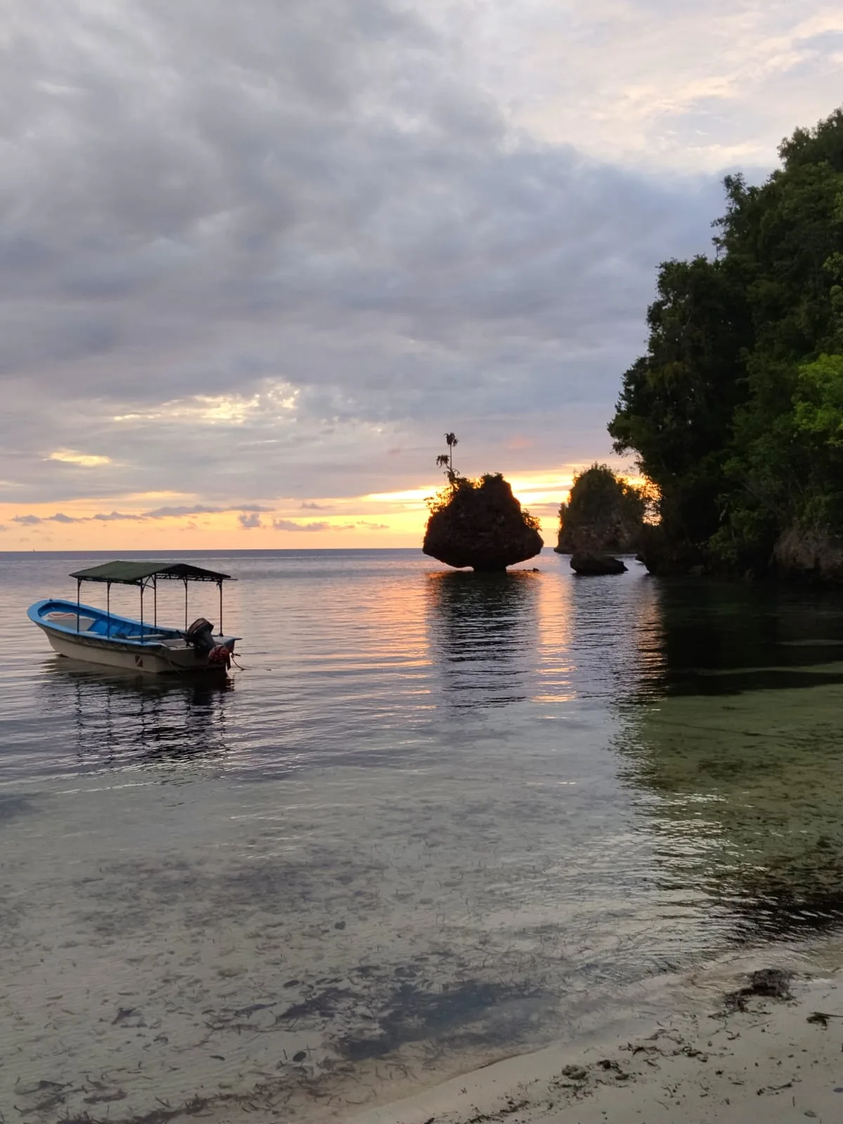 A small canopied boat anchored near mushroom-shaped rock outcrops in calm water at sunset, with forested cliffs on the right
