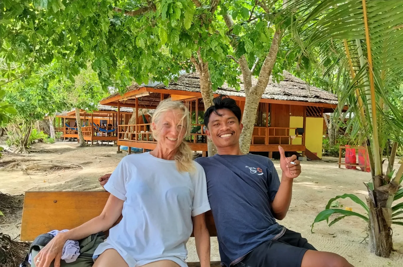 Two people sit on a bench smiling under a large tree, with wooden thatched-roof cottages in the background