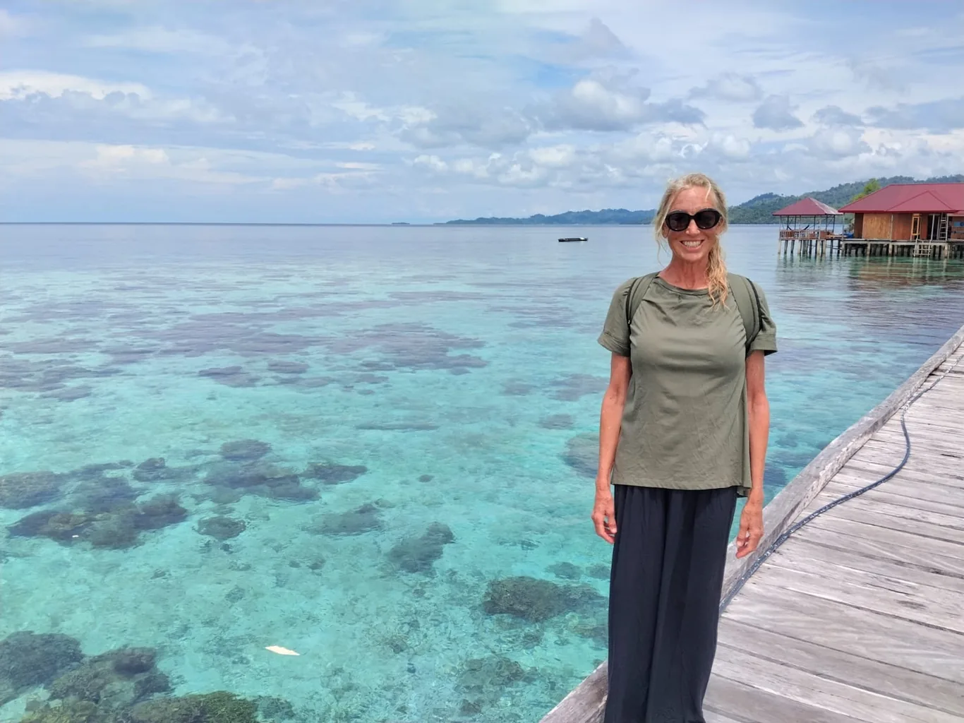 A woman in sunglasses stands on a wooden jetty over clear turquoise water with visible coral, an overwater cottage in the background