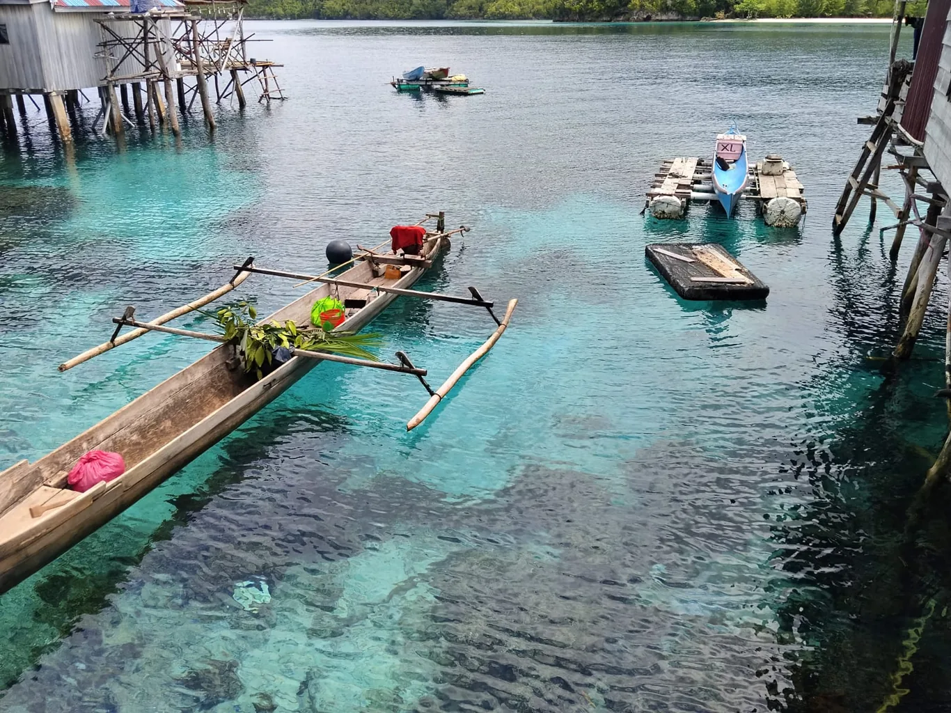 Wooden outrigger canoe with palm leaves floats over clear turquoise water near stilt houses and a small moored motorboat
