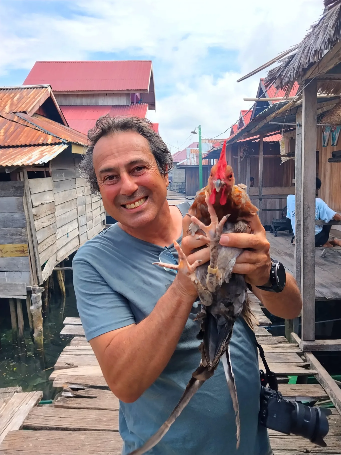 A smiling man in a blue shirt holds a rooster by its legs while standing on a wooden walkway between stilted buildings