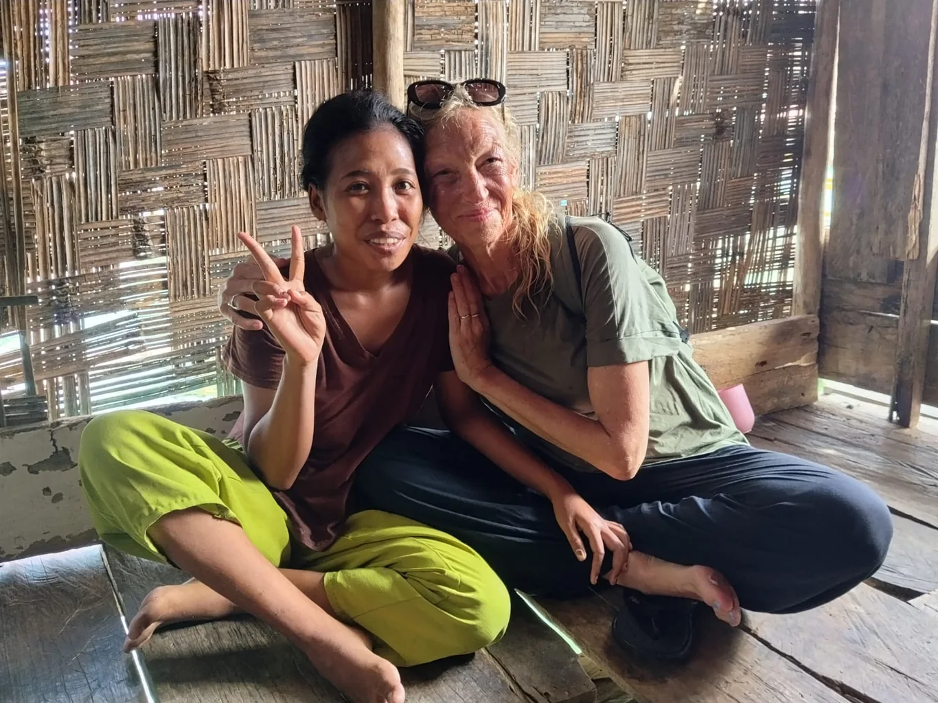 Two women sit together smiling on a wooden floor, one giving a peace sign, in front of a woven bamboo wall