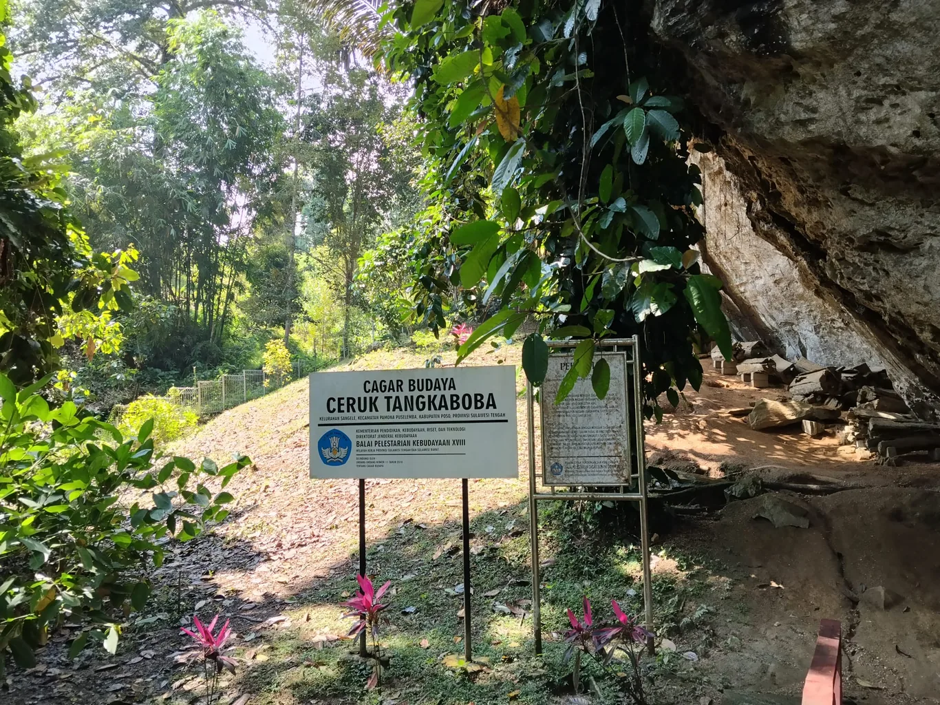 A signboard reading 'Cagar Budaya Ceruk Tangkaboba' stands near a rock overhang entrance surrounded by forest and pink flowers