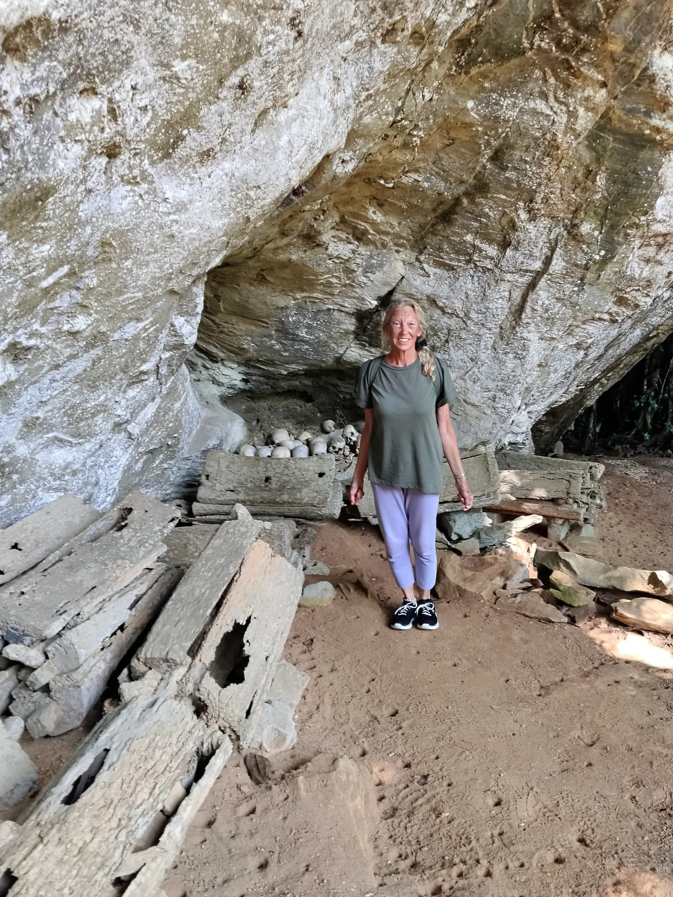 A woman stands smiling inside a rock shelter beside wooden coffin fragments and skulls arranged on the ground
