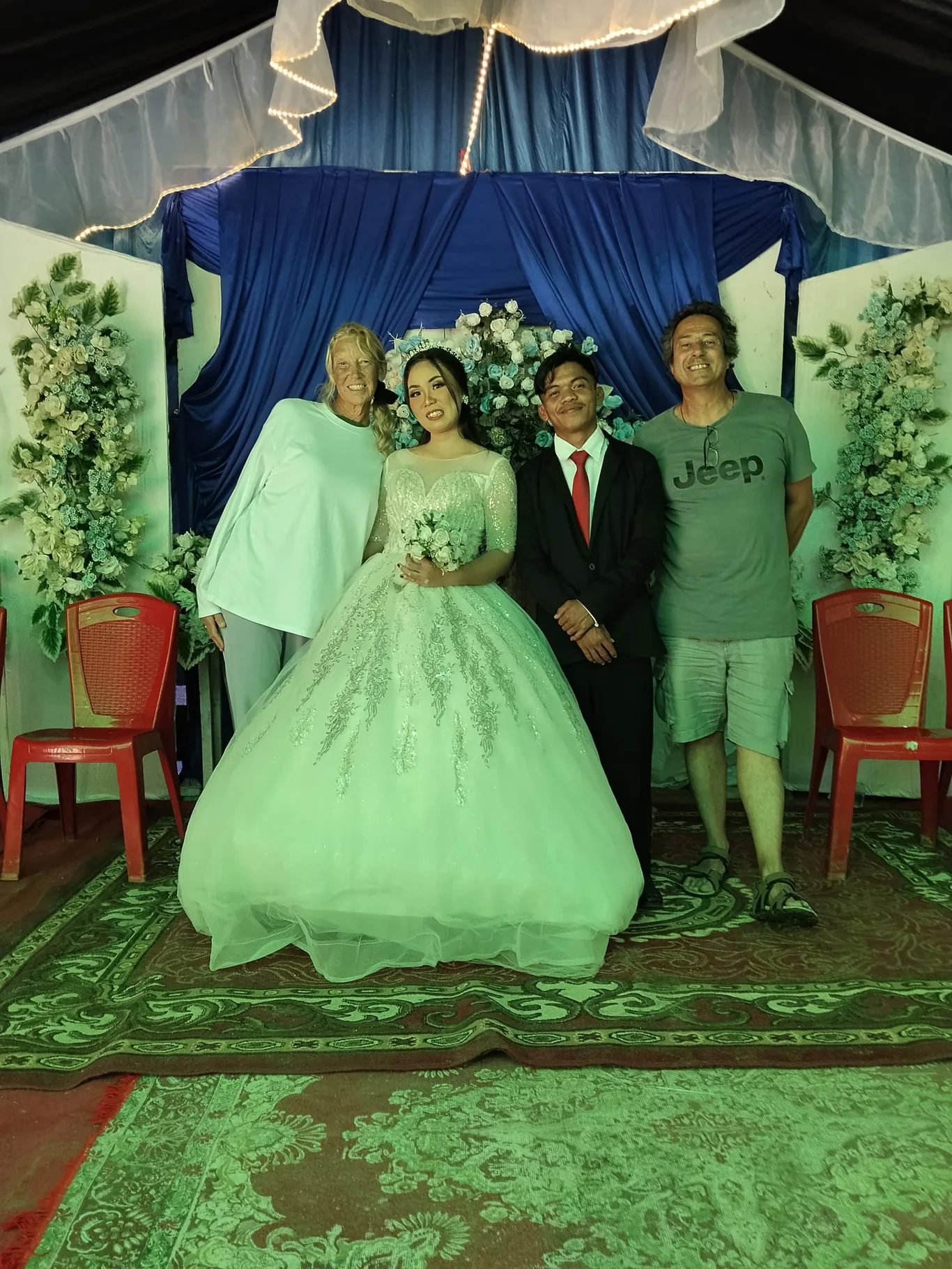 Four people pose at a decorated stage with a bride in a white gown, flanked by floral arrangements and blue drapery