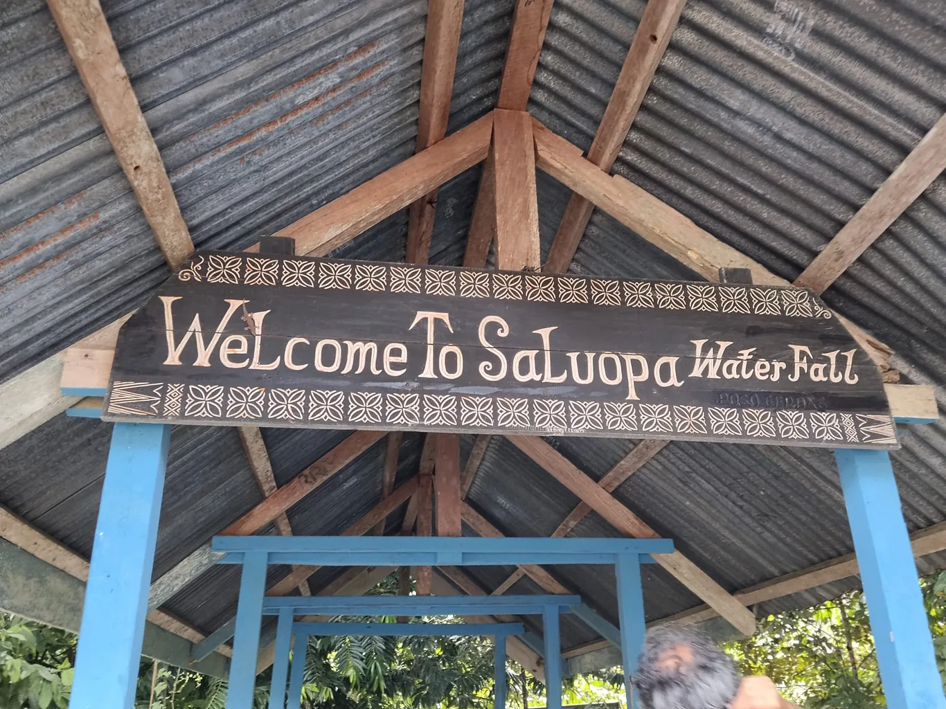 A carved wooden sign reading 'Welcome to Saluopa Waterfall' hangs beneath a corrugated metal roof supported by wooden beams and blue pillars