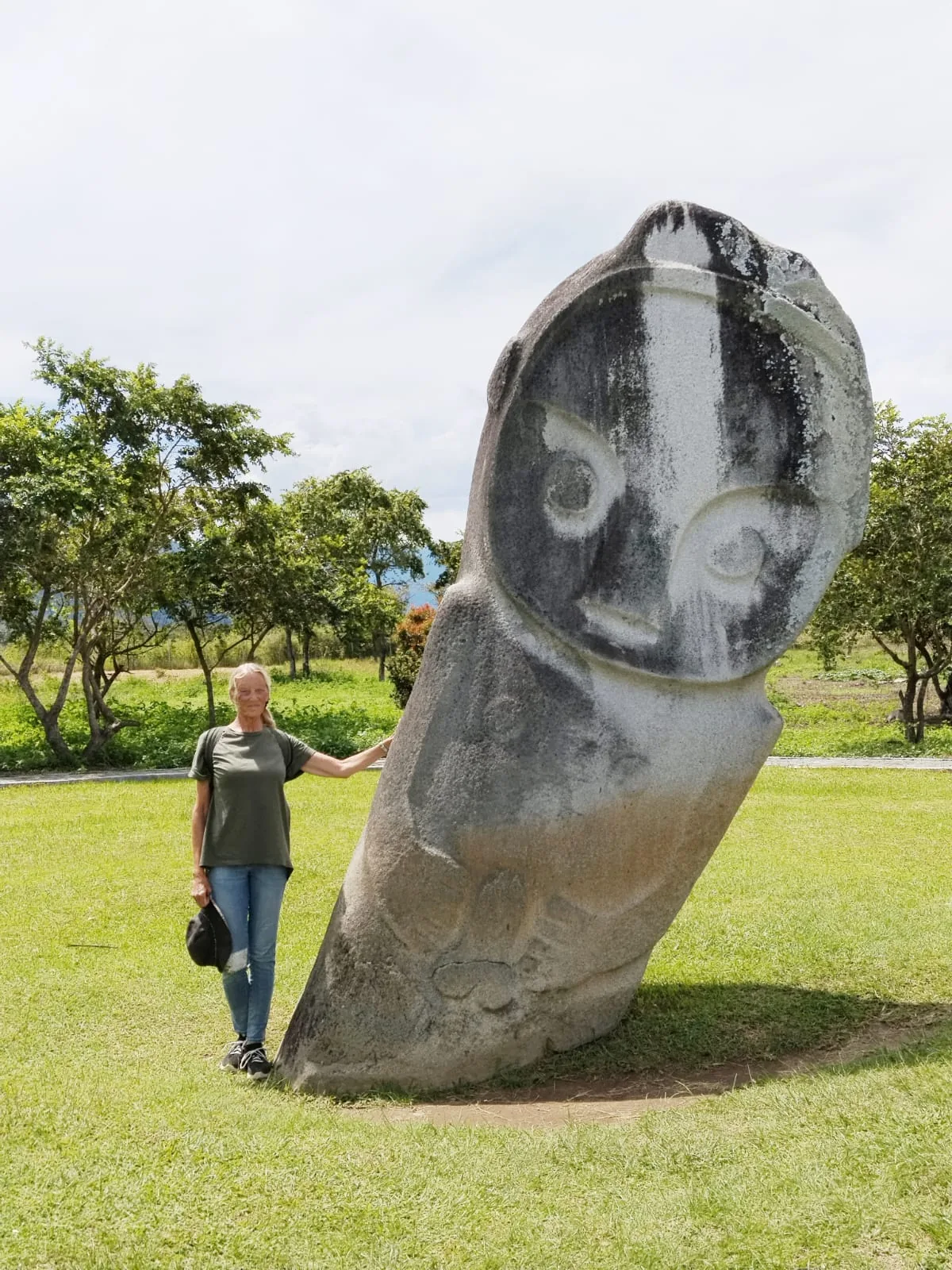 A woman stands beside a large stone statue carved with a face, resembling an owl, set in a grassy field