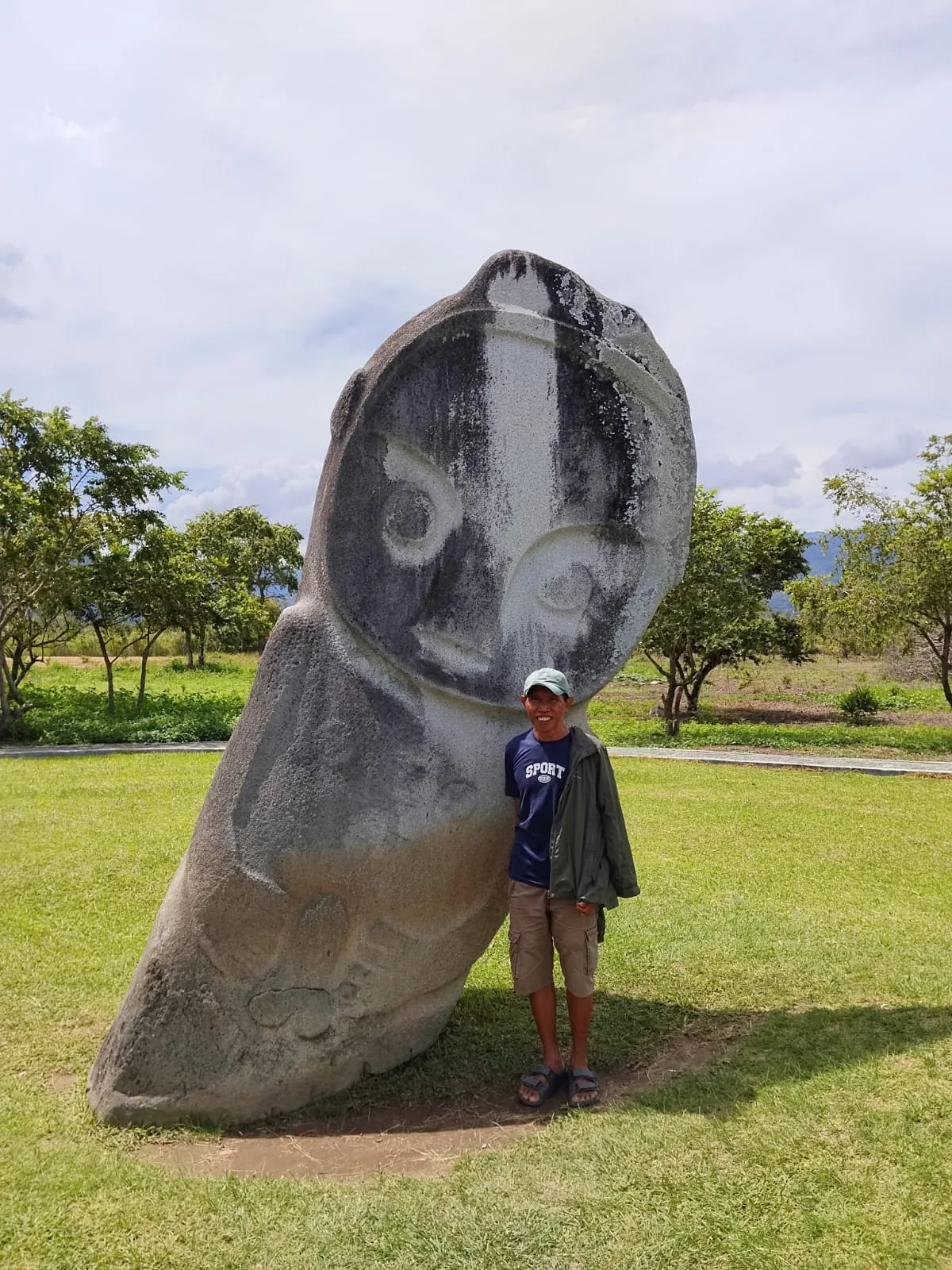 A young man in a cap smiles beside a large carved stone figure with painted facial features, standing on open grass