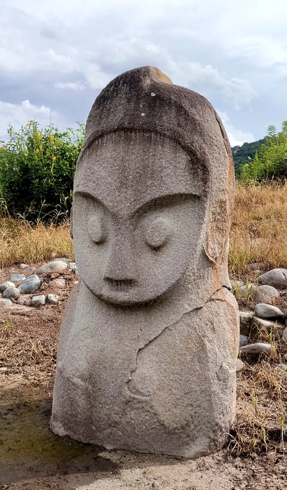 A weathered stone statue with a simple carved face stands in dry grass, surrounded by scattered rocks and green shrubs