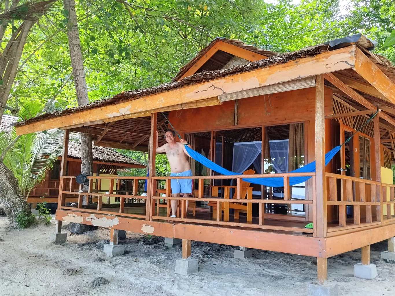A shirtless guest stands on a wooden bungalow's porch beside a blue hammock, with a shaded sandy path below