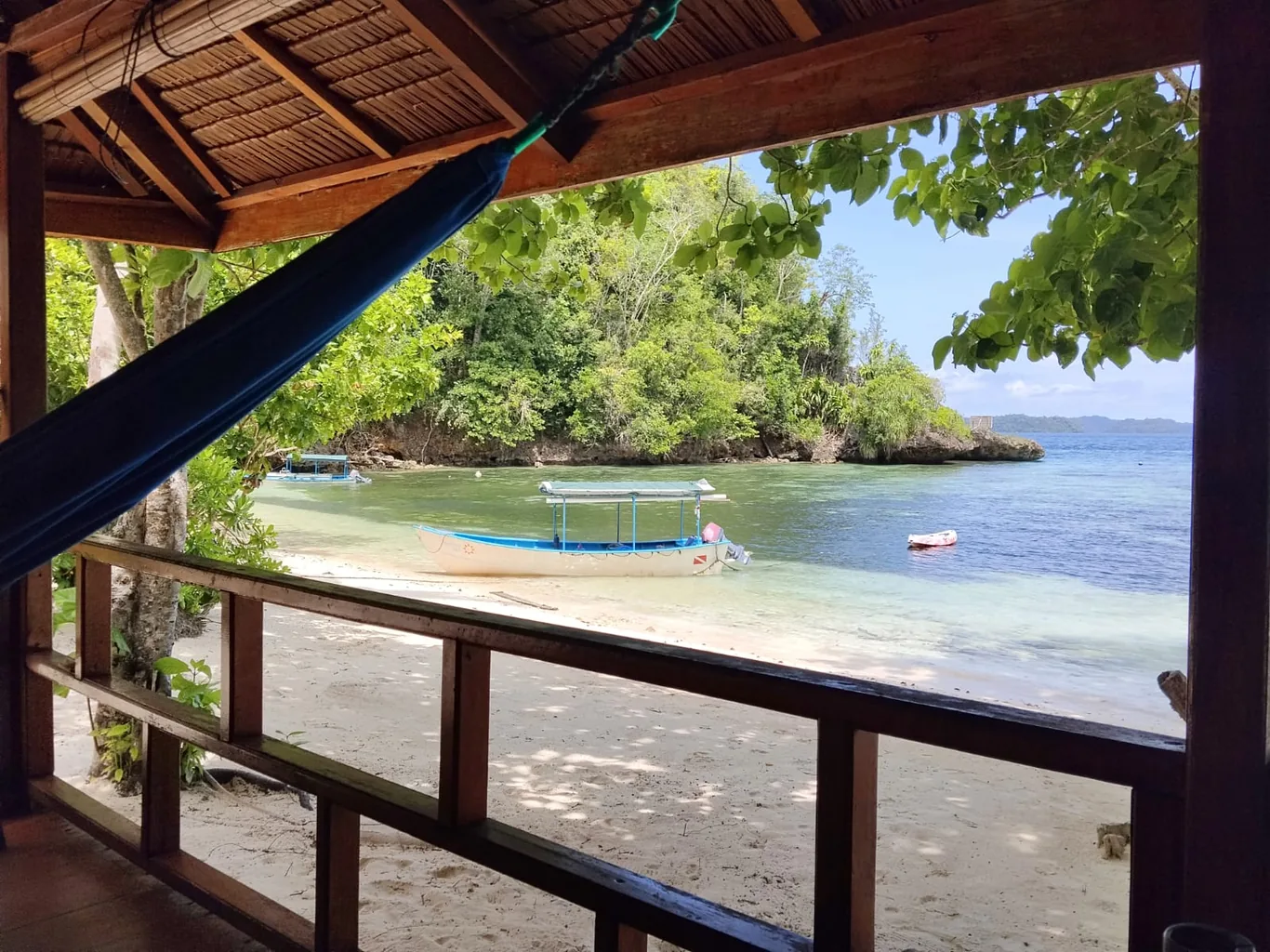 View from a wooden thatched-roof porch with a hanging hammock overlooking a beached wooden boat on a sandy shore with turquoise water
