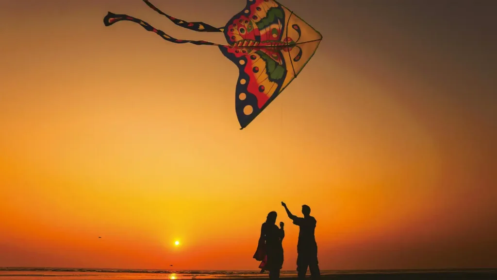Silhouettes of two people flying a large colorful kite on a beach at sunset with the sun low on the horizon