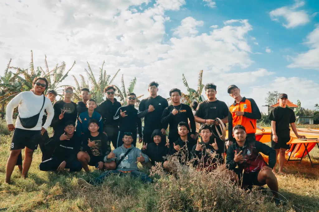 Group of young men posing together outdoors in a grassy field with banana trees and a partly cloudy sky behind them