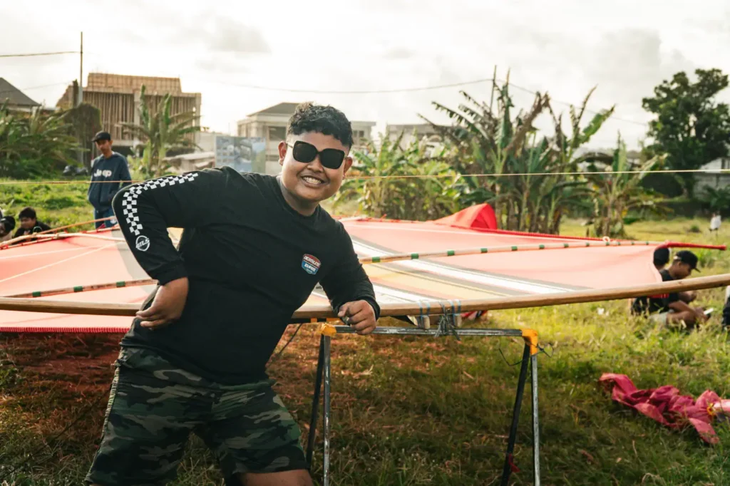 A man in sunglasses smiles while posing beside a large traditional kite frame laid out on grass