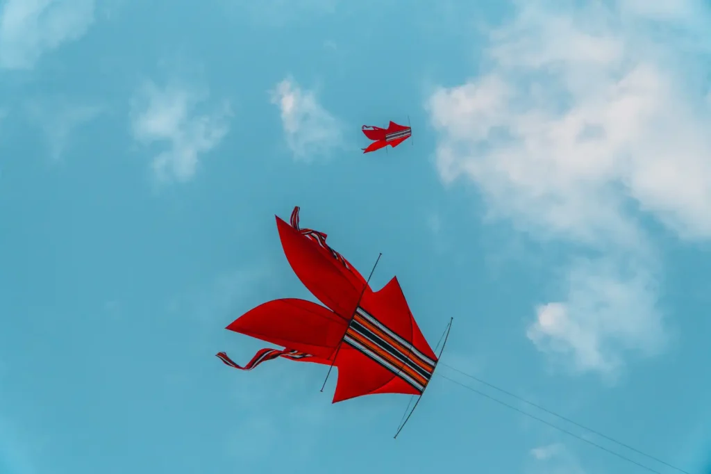 Two red traditional kites with long ribbon tails flying against a blue sky with scattered white clouds