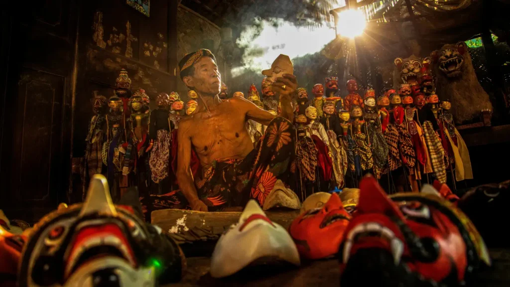 An elderly bare-chested man carves a wooden puppet, surrounded by rows of traditional masks and puppet figures lit by backlight