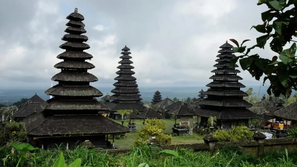 Multi-tiered thatched-roof pagoda towers rise above a walled temple complex with stone shrines and lawns under a cloudy sky