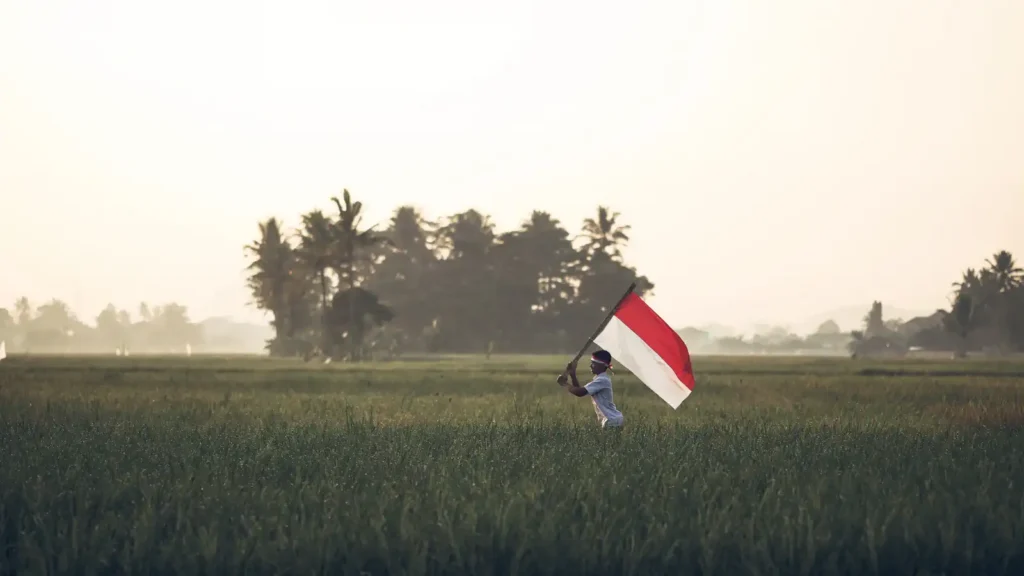 A person runs through a green rice field waving a red and white flag, trees and hazy sky in the background
