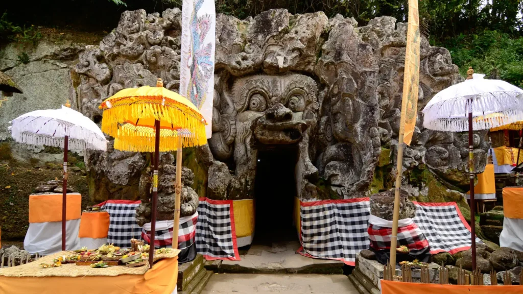 Carved demon-face entrance of the Goa Gajah cave with ceremonial umbrellas and offerings