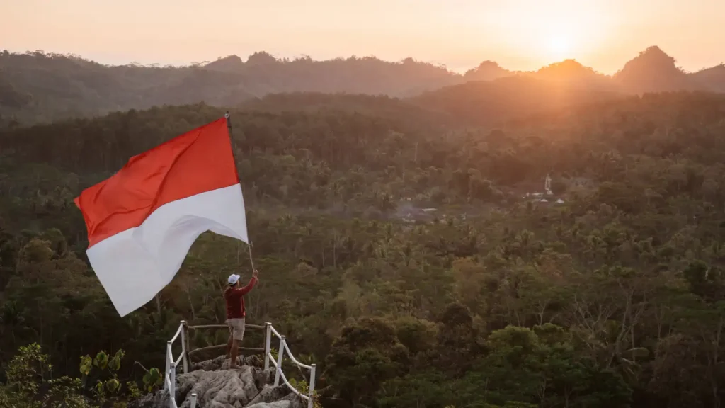 A visitor raising the Indonesian flag at a hilltop viewpoint above forest at sunrise