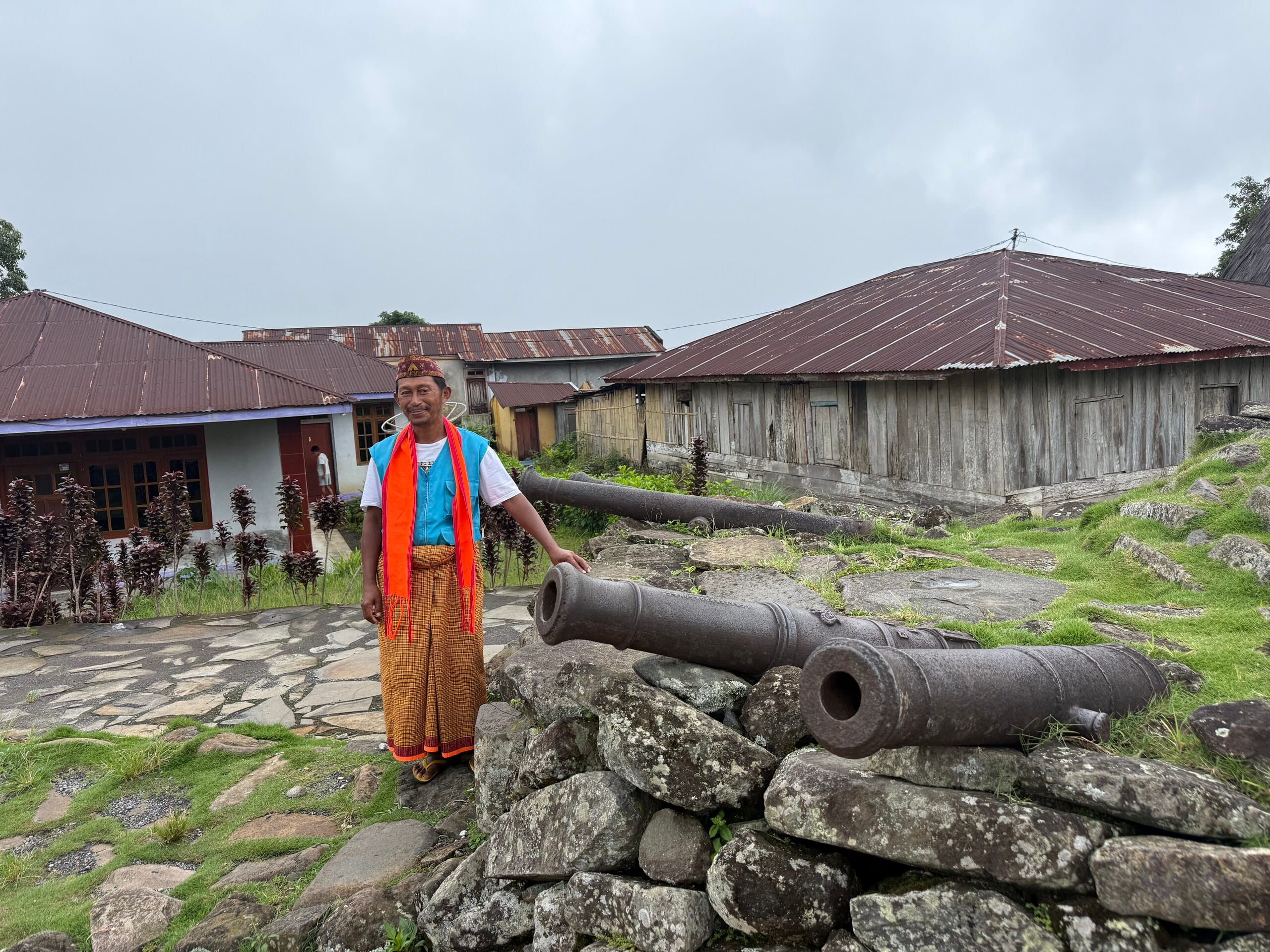 Dean in Manggarai dress beside two old iron cannons on a stone wall at Compang Todo
